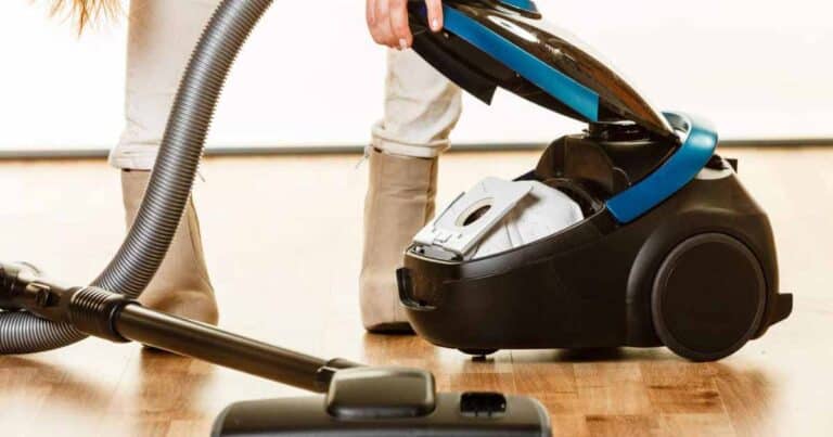 A person wearing beige boots is changing the dust bag of a black and blue canister vacuum cleaner on a wooden floor. The vacuum hose is extended, ensuring every corner is reached, while the integrated HEPA filter promises to capture even the finest dust particles.