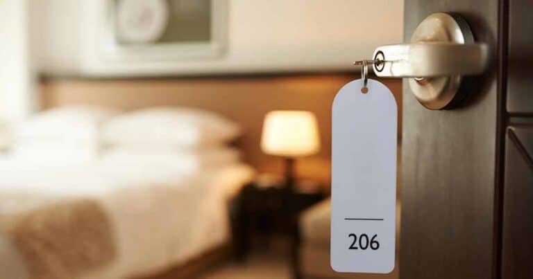 A hotel room door is slightly open, revealing a cozy, allergy-friendly interior with a neatly made bed and a lit bedside lamp. A key is inserted in the door lock with a white tag displaying the number 206.