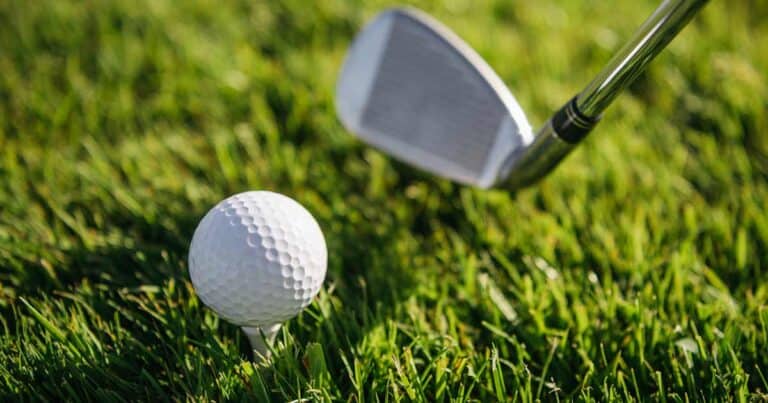 A close-up of a golf club about to hit a white golf ball sitting on a tee, both positioned on green grass.