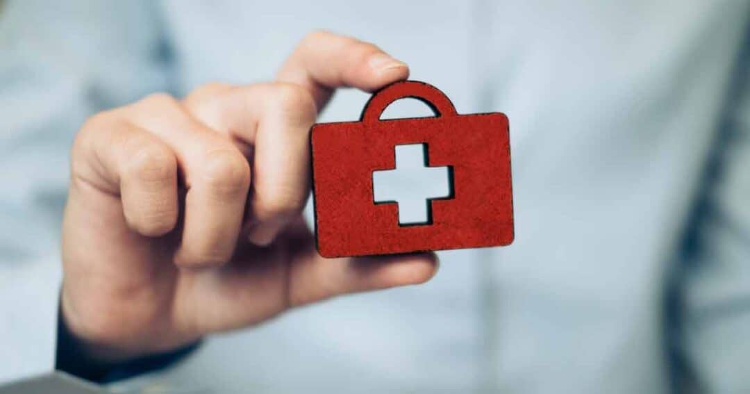 A person holding a small red first aid kit symbol with a white cross on it. The background is blurred, highlighting the focus on the hand and the symbol.