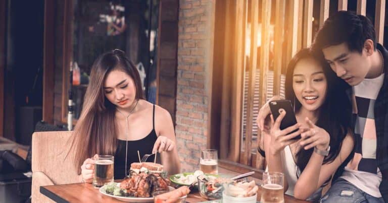 Three people are seated at a restaurant table. A woman on the left is focused on her meal, carefully selecting dishes due to her food allergies. The other two are smiling at a phone. Various dishes and drinks are on the table. The setting is warm and inviting, perfect for eating out.