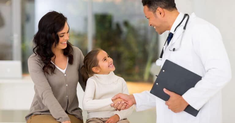 Doctor standing up facing a mom and her tween child. He is shaking the child's hand and they are all smiling.