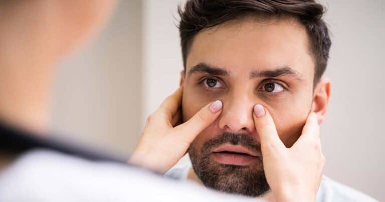 A person is having their eyes inspected by a healthcare professional, who gently places their fingers around the person's eyes, checking for signs of sinus pressure. The background is blurred and indistinct.