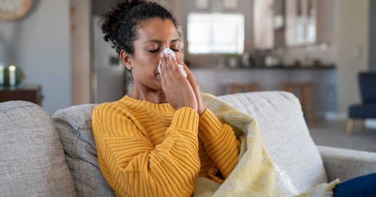 Woman with stuffed nasal passages sitting on her couch blowing her nose