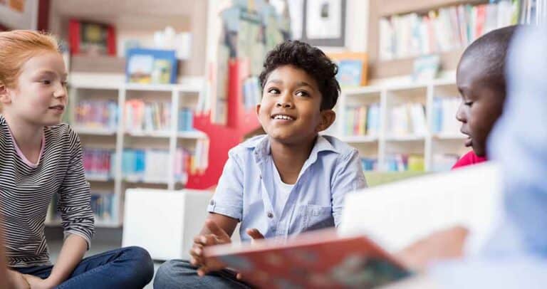 Multiethnic group of kids sitting on floor in circle around the teacher and listening a story. Discussion group of children inaa library talking to woman. Smiling hispanic boy in elementary school.