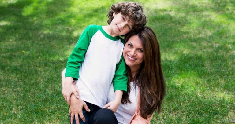 A joyful woman with long hair sits on the grass, embracing a young boy with curly hair wearing a green and white shirt. Both are smiling warmly at the camera. The background features a lush green lawn.
