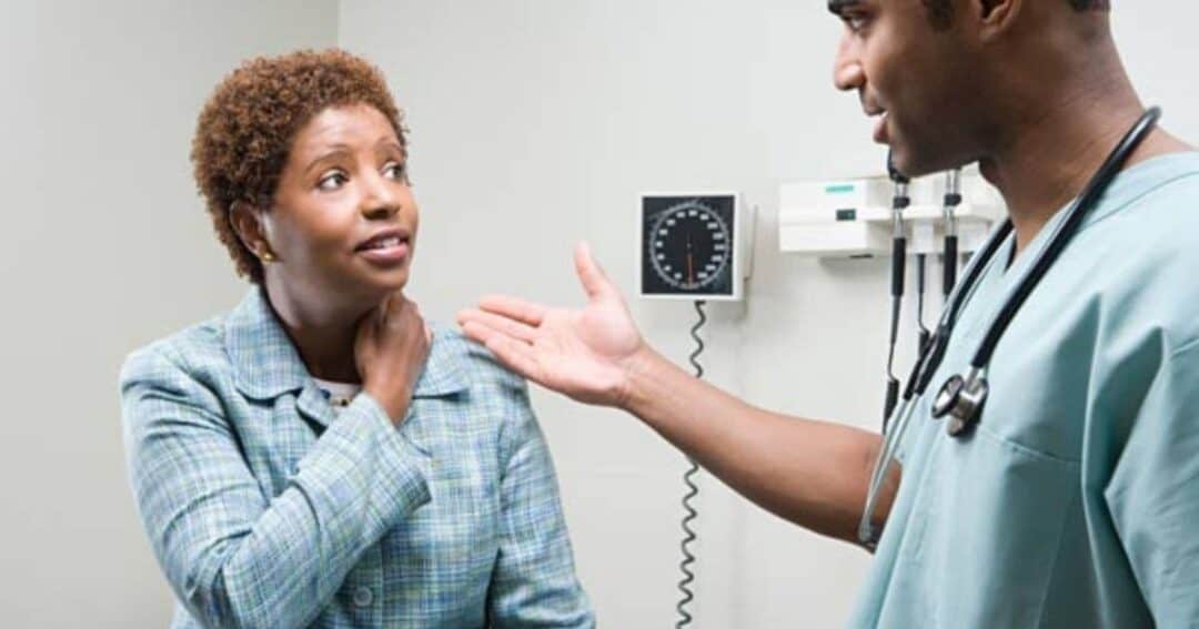 A doctor in medical scrubs is speaking to a patient in an examination room. The patient, wearing a blue jacket, appears concerned and touches her neck. A blood pressure monitor is visible on the wall in the background.