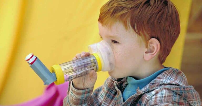 A young child with red hair is using an inhaler with a spacer. The child is wearing a plaid shirt and is focused on the device. The background is softly blurred in yellow tones.
