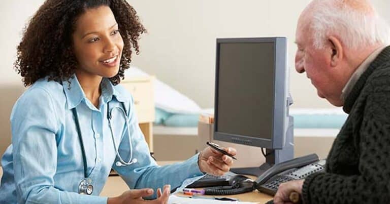 A healthcare professional is sitting at a desk, talking to an elderly man. The professional is holding a pen and wearing a stethoscope. They both appear engaged in conversation, with a computer monitor and telephone visible on the desk.