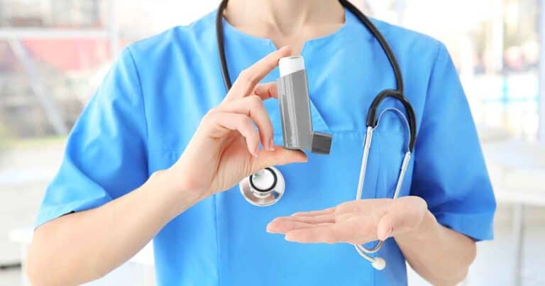 A healthcare professional in a blue uniform with a stethoscope around the neck is holding an asthma inhaler. The focus is on the inhaler, which is gray with a white top, against a blurred background.