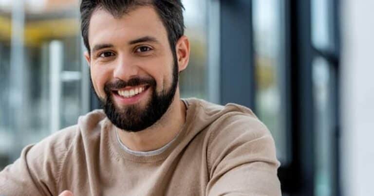 A man with a beard and short hair is smiling at the camera. He is wearing a beige sweater and is indoors, with blurred glass windows in the background.