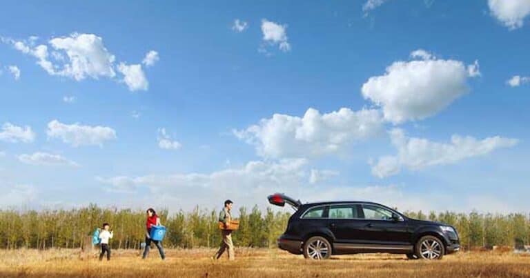 A family of four is walking in a grassy field towards a black SUV with an open trunk, carrying camping gear. The sky is blue with scattered clouds, and a line of trees is visible in the background.