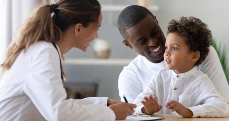 A female doctor in a white coat smiles while talking to a father and his young son. The father is also smiling at the child, who seems attentive. They are sitting at a table in a bright room.