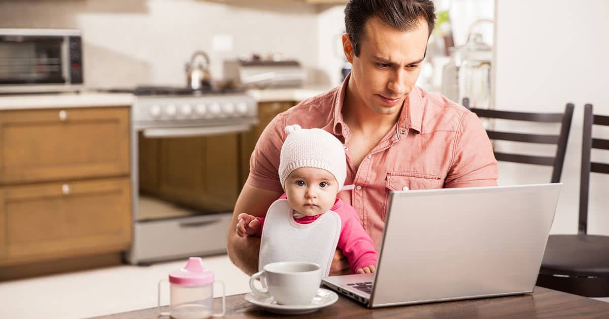 A man in a pink shirt sits at a kitchen table holding a baby wearing a pink hat and bib. The man is focused on a laptop. A cup and a baby bottle with a pink lid are on the table. The kitchen is in the background.