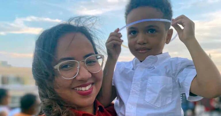 A woman with glasses smiles while holding a young boy. The boy is adjusting a clear visor on his head. They are outdoors, with a partly cloudy sky in the background.