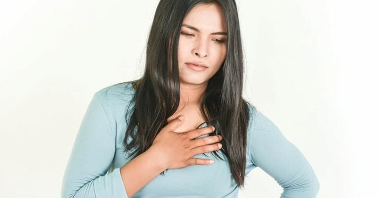 A woman with long dark hair wearing a light blue shirt holds her chest with one hand, looking slightly discomforted. She stands against a plain white background.