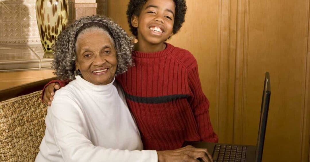 A smiling elderly woman with gray hair and a young boy in a red sweater sit together at a laptop. The boy has his arm around the woman, and they are in a cozy room with wooden cabinets.