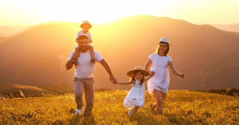 A family of four enjoys a sunny day in a grassy field, with mountains in the background. The father carries a child on his shoulders, while the mother and daughter walk beside them, all smiling. Everyone is dressed in light, summer clothing.