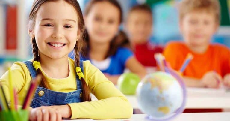 A smiling girl with braided hair sits at a desk in a classroom, wearing a yellow shirt and blue overalls. A small globe and colored pencils are on the desk. Other children are visible in the background.