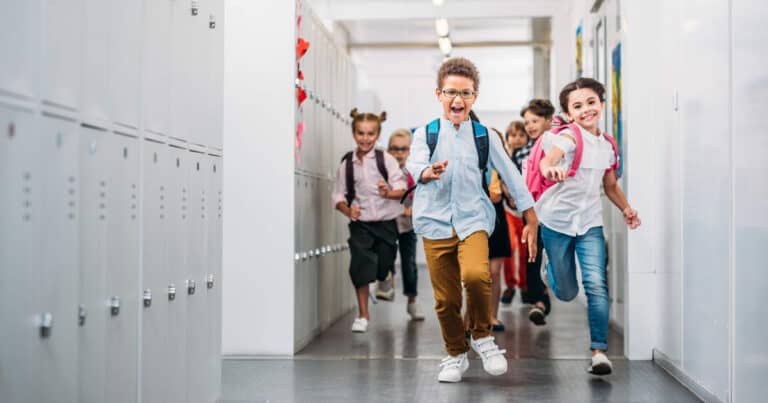 A group of excited children with backpacks run down a school hallway lined with lockers. They are laughing and appear happy as they head towards the camera.