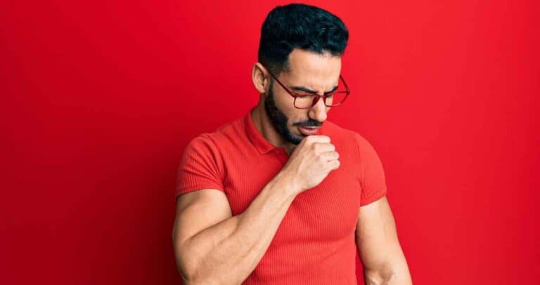 A man with a beard and glasses wearing a red polo shirt stands against a matching red background. He is looking down with his hand near his mouth, appearing thoughtful or contemplative.