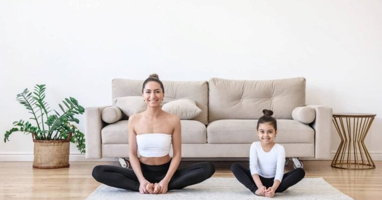 A woman and a young girl are sitting cross-legged on a gray mat in a living room. Both are smiling and wearing white tops and black leggings. Behind them, there is a beige couch and a potted plant on a wooden floor.