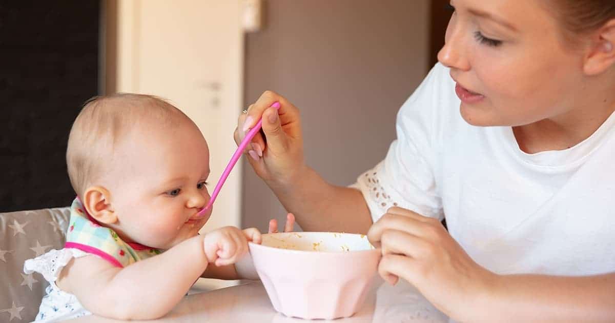A woman is feeding a baby with a pink spoon at a table. The baby is wearing a bib and is focused on the spoon, while the woman looks at the baby with a gentle expression. A pink bowl is placed in front of them.