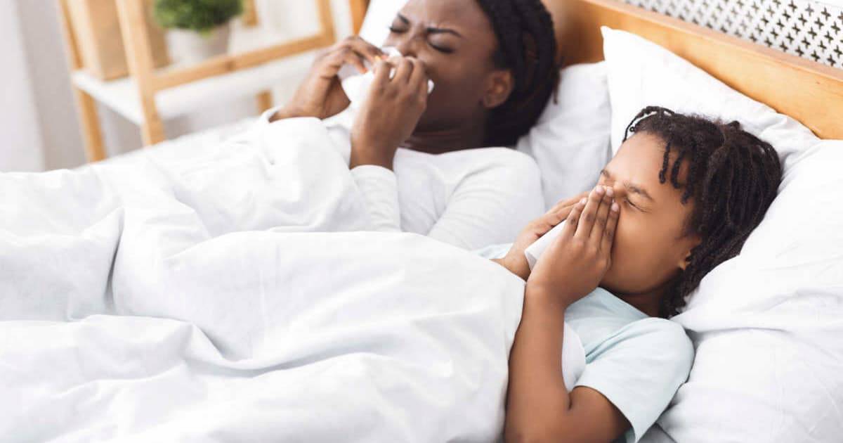 A child and an adult lie in bed under a white blanket, both holding tissues to their noses, suggesting they might have a cold. The room has light-colored walls and a wooden shelf in the background.