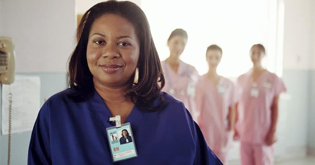 A nurse in blue scrubs stands confidently in the foreground, wearing an ID badge. Three nurses in pink scrubs stand behind her, slightly out of focus. They are in a brightly lit hospital hallway.