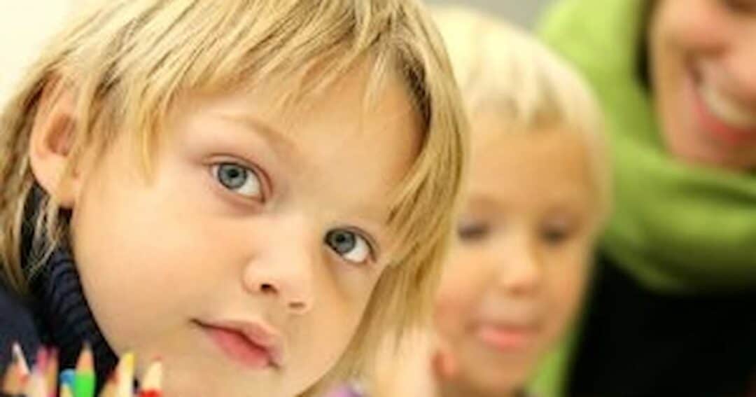 A close-up of a child with blond hair looking at the camera, surrounded by colorful pencils. Another child and an adult with a green scarf are blurred in the background, smiling.