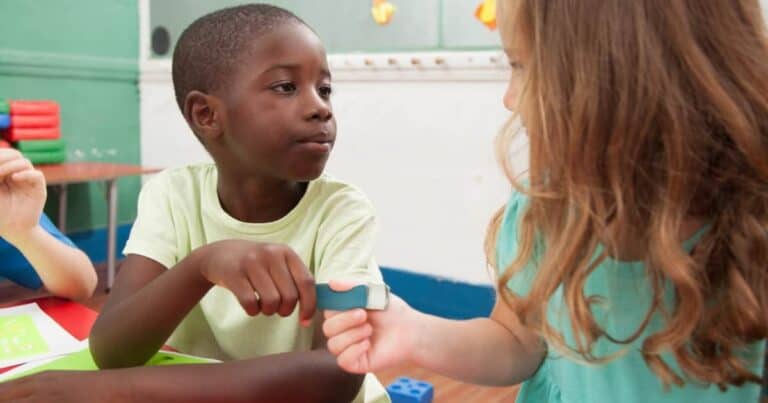 A young boy and girl sitting at a table in a classroom, engaged in an activity. The boy is holding a tool, and they are focused on a task together. The classroom is colorful, with green and blue walls and toys in the background.