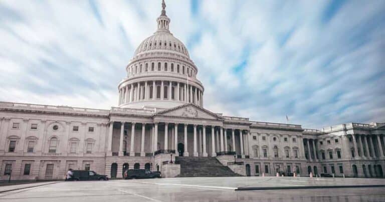 The image shows the United States Capitol building with its large dome and neoclassical architecture. The sky above is partly cloudy, and a few vehicles are parked nearby.