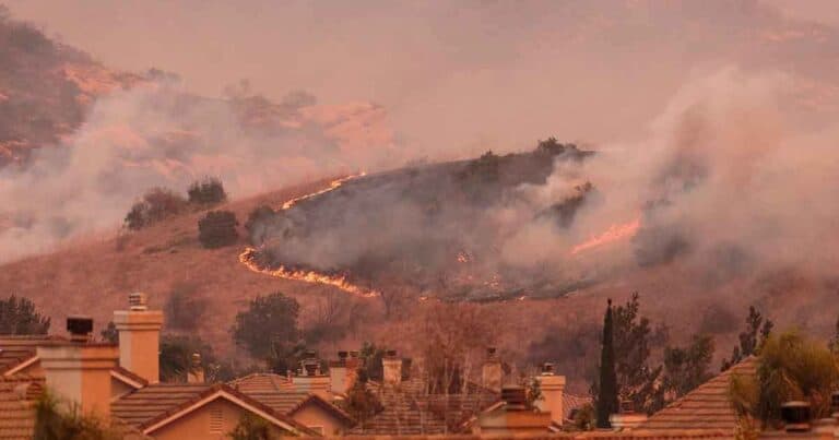A hillside is engulfed in smoke and flames during a wildfire, with visible fire lines along the dry grass. Roofs of houses are seen in the foreground, under a hazy, smoky sky.