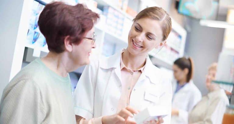 A pharmacist in a white coat smiles and assists an elderly woman with a medication package in a pharmacy. Shelves of pharmaceutical products are visible in the background, with another pharmacist working nearby.