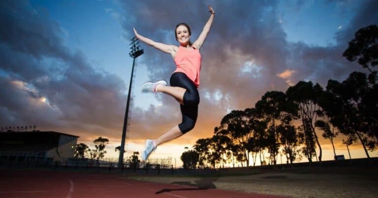 A woman in athletic wear jumps joyfully on a track at sunset. The sky is filled with dramatic clouds, and silhouetted trees line the horizon.