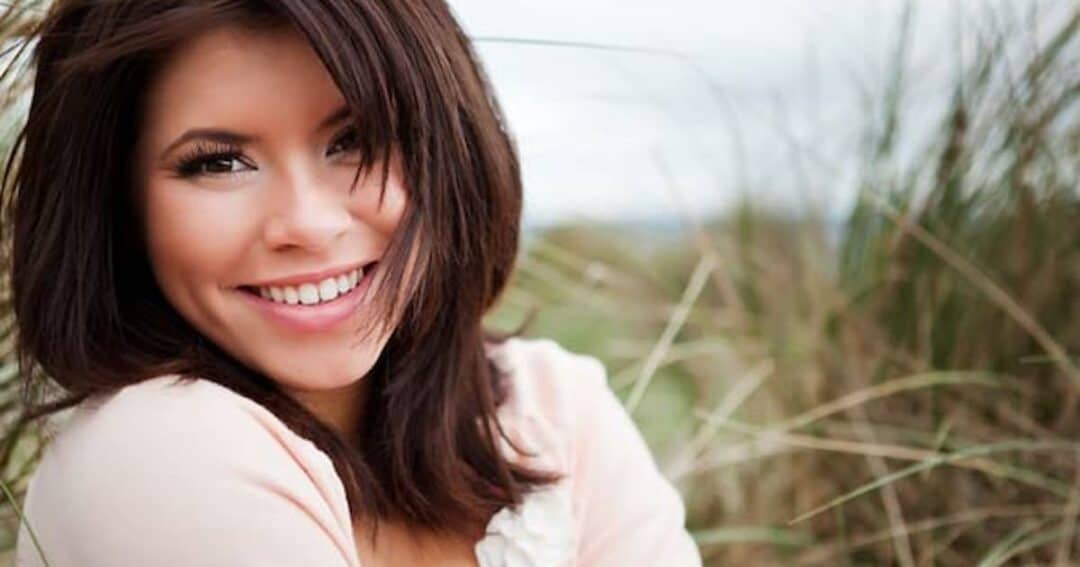 A woman with dark hair smiles warmly at the camera, standing outdoors amidst tall grass. She is wearing a light-colored top, and the background is slightly blurred, suggesting a breezy, natural setting.