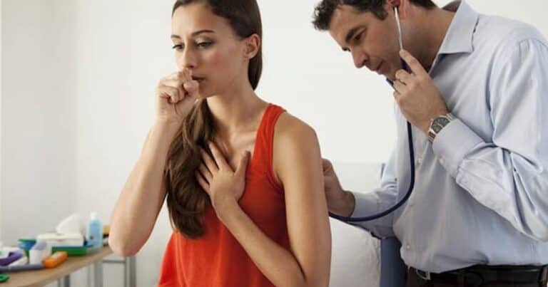 A woman in a red tank top coughs into her hand while a doctor listens to her back using a stethoscope in a medical office.