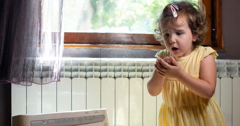 A young girl in a yellow dress stands by a radiator and holds her hands to her face as she sneezes, with sunlight streaming through a window, illuminating dust particles in the air.