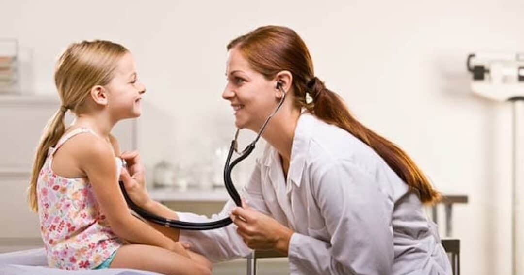 A female doctor in a white coat uses a stethoscope to examine a smiling young girl sitting on an examination table in a medical office.