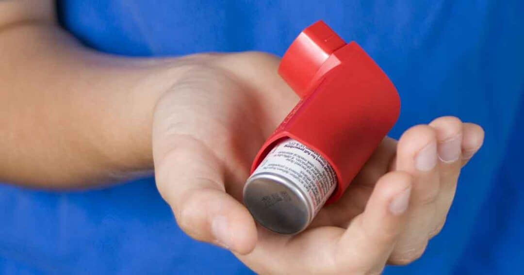 A person holds a red asthma inhaler in their hand against a blue background.