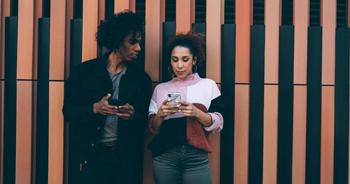 Two people stand against a patterned wall, each holding a smartphone. The person on the left, wearing a dark jacket, looks at the other's phone. The person on the right wears a colorful sweater and focuses on their screen.