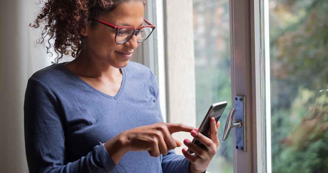 A person with curly hair and glasses is smiling while using a smartphone near a window. They are wearing a blue shirt and appear focused on the device. Natural light from outside illuminates the scene.