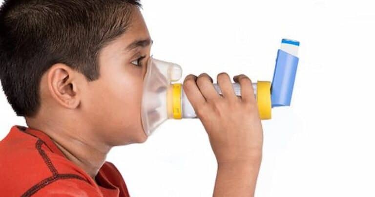 A child wearing a red shirt uses an inhaler with a yellow spacer and a transparent mask, against a white background.