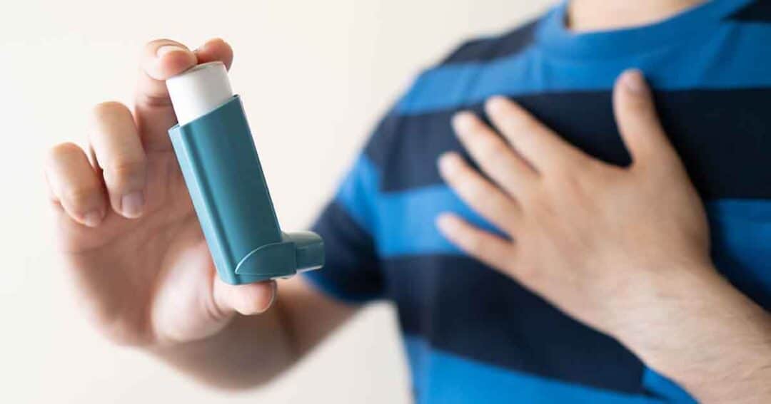 A person in a striped shirt holds a blue asthma inhaler in one hand while touching their chest with the other hand, indicating readiness to use the inhaler.
