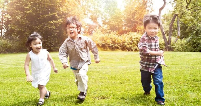 Three children joyfully running across a grassy field. The sun is shining brightly, and trees are visible in the background, creating a warm, cheerful atmosphere. The kids look happy and playful.