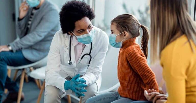A doctor in a white coat and mask is kneeling and talking to a young girl wearing a mask and orange sweater. The girl is sitting next to an adult, also wearing a mask, in a waiting area with other people seated in the background.