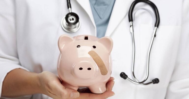 A doctor wearing a white coat holds a pink piggy bank with a band-aid on its side. A stethoscope is draped around the doctor's neck, symbolizing healthcare costs or savings.