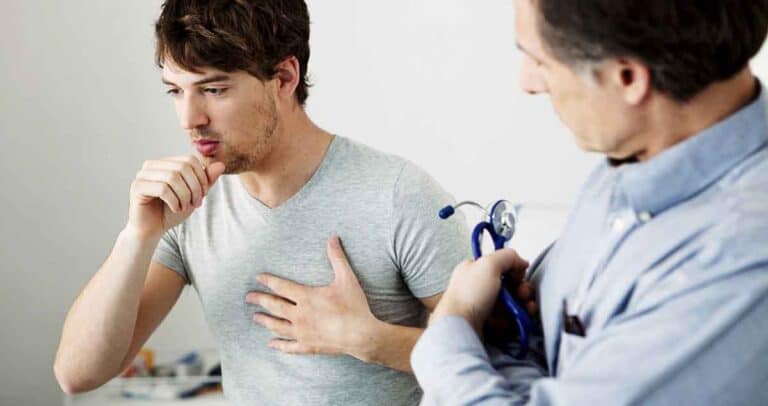 A young man wearing a gray T-shirt is coughing and touching his chest. He is at a medical consultation with a doctor, who is observing him and holding a stethoscope.