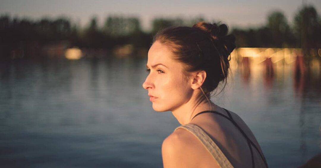 A woman with her hair in a bun is gazing thoughtfully to the side while seated by a calm lake during sunset. The background features a blurred view of trees and soft evening light.