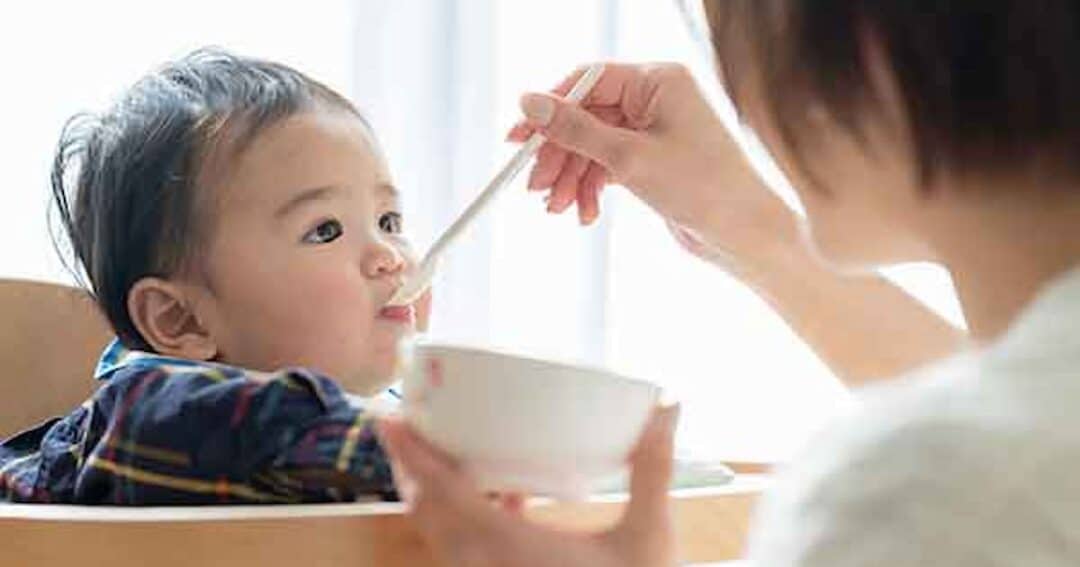 A baby sits in a high chair, being fed by an adult. The baby looks curiously at the spoon approaching its mouth. The adult holds a white bowl while gently feeding the baby in a bright, softly lit room.